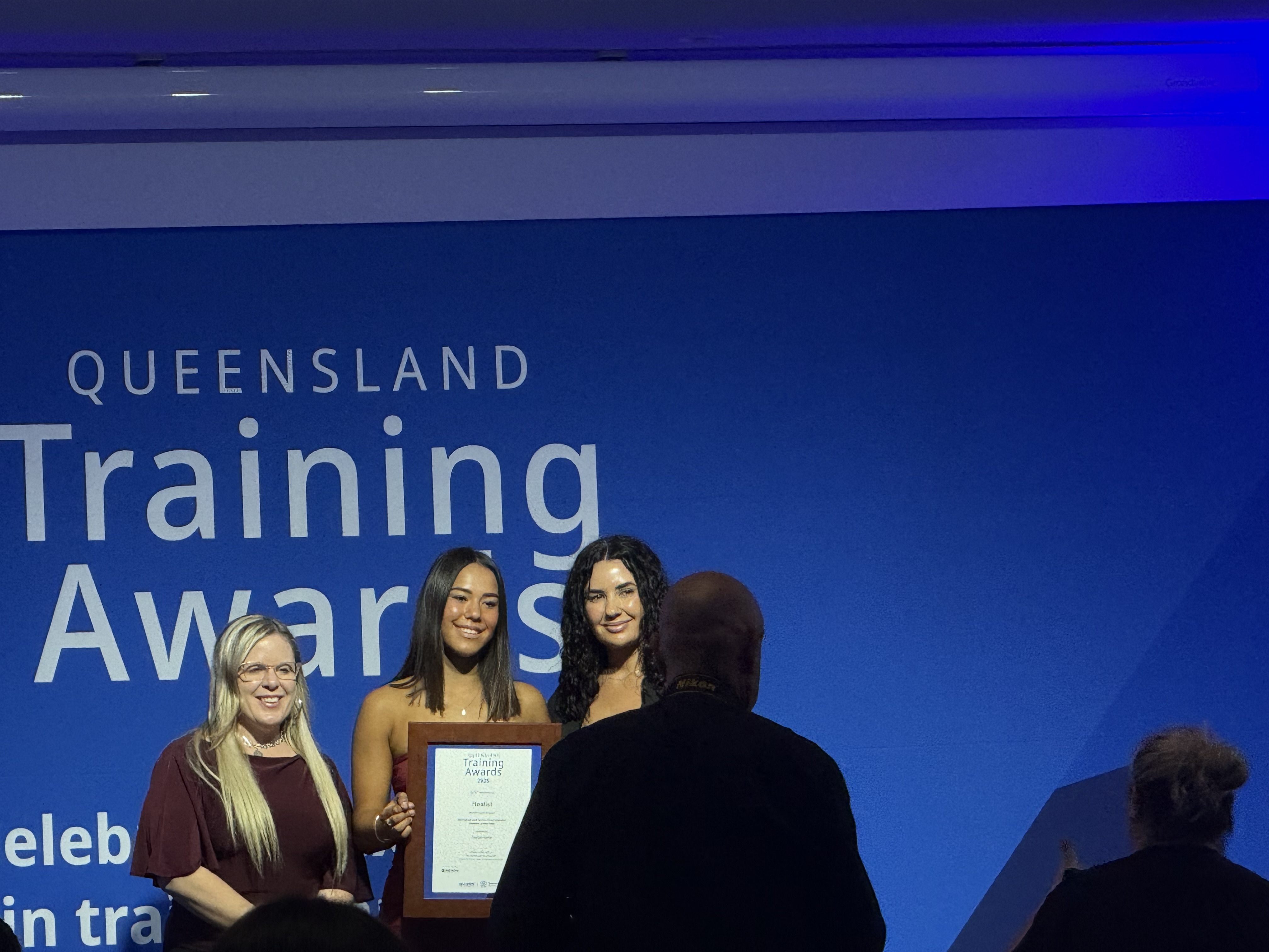 A trainee receiving a framed Queensland Training Awards certificate on stage in front of the Queensland Training Awards backdrop