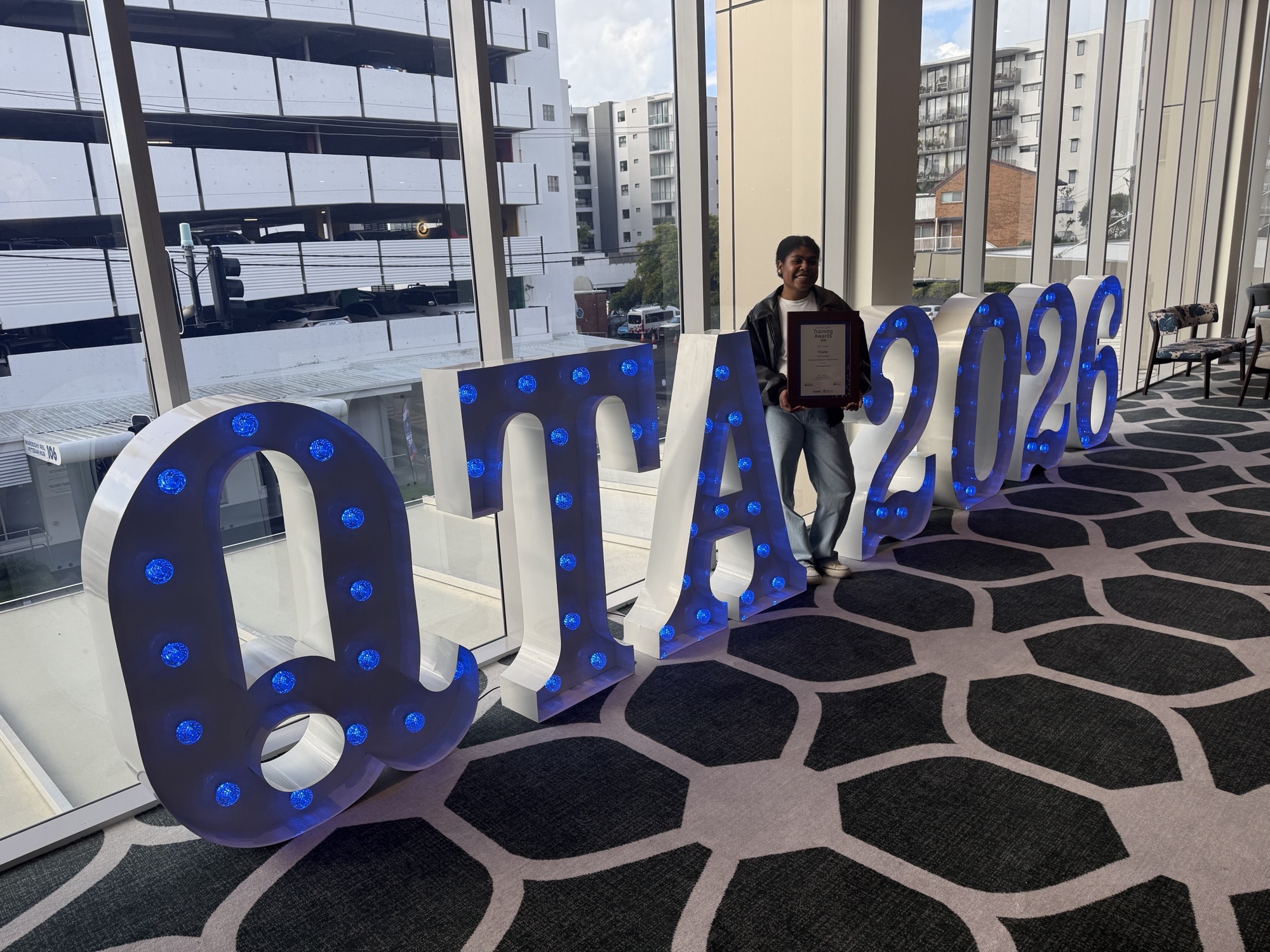 Genevieve Jimmy holding her framed Queensland Training Awards finalist certificate beside the illuminated QTA 2026 letters at the awards venue